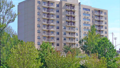 A tall concrete building with many balconies and trees in front near a road. 