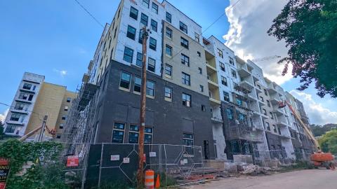 A photo of a large brick and stucco apartment complex that's taken shape under blue skies beside green trees.