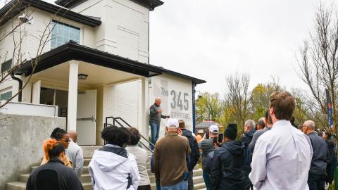 A photo of a group of people touring a large white building that's affordable housing on a rainy day.