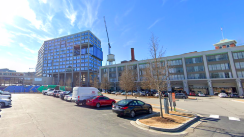 A photo of a parking lot surrounded by a variety of tall buildings under blue skies. 