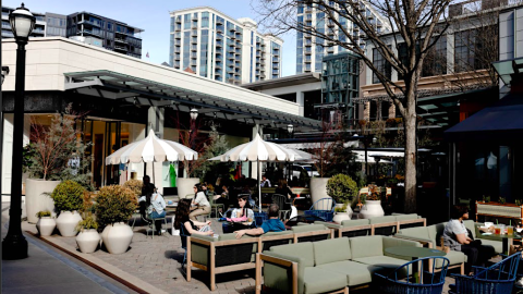 An image of a shopping district with many people on patios under a blue sky.