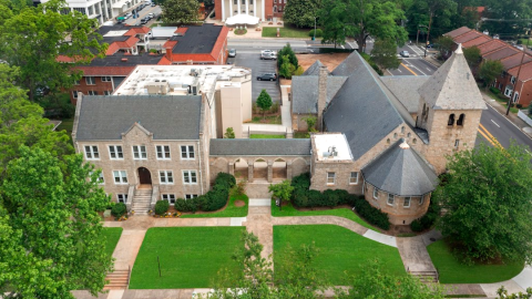 A photo of a large old stone building and chapel on a corner in a city with lawns in front and white-painted offices inside.