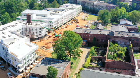 An aerial image of a collection of old brick buildings next to a water tower and stack of white apartments near red dirt.