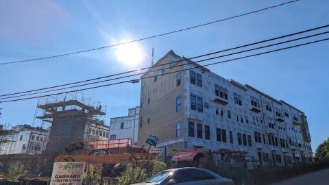 An image of a long row of townhomes under blue skies near a busy street, with rooftop patios above. 