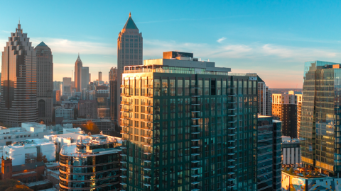 A photo of a large green-clad and glass building in Midtown Atlanta amid many more skyrise under a sunrise.