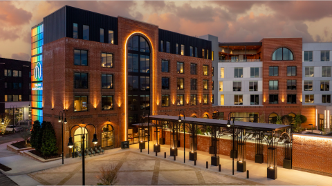 A photo of a new brick-clad hotel with modern interiors and a bar on the roof and a pool behind it, in a new small town. 