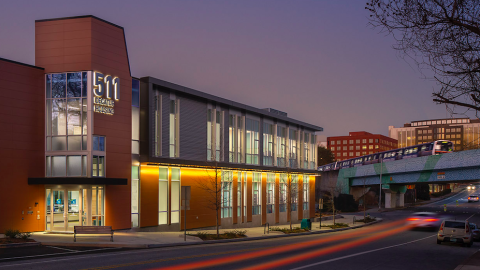 A photo of a modern two-story new building with brown and silver panels on the exterior, many windows, and art and classrooms in the white-painted interior. 
