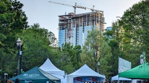 A large tower of glass and concrete shown next to a park with a large arts festival ongoing.