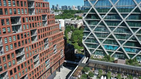 A drone photo of a large new development with three towers over a large city park under blue skies, with a city skyline in the distance.