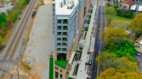 A large wedge-shaped apartment building next to railroad tracks and streets in Atlanta and many trees, with a pool on the roof. 
